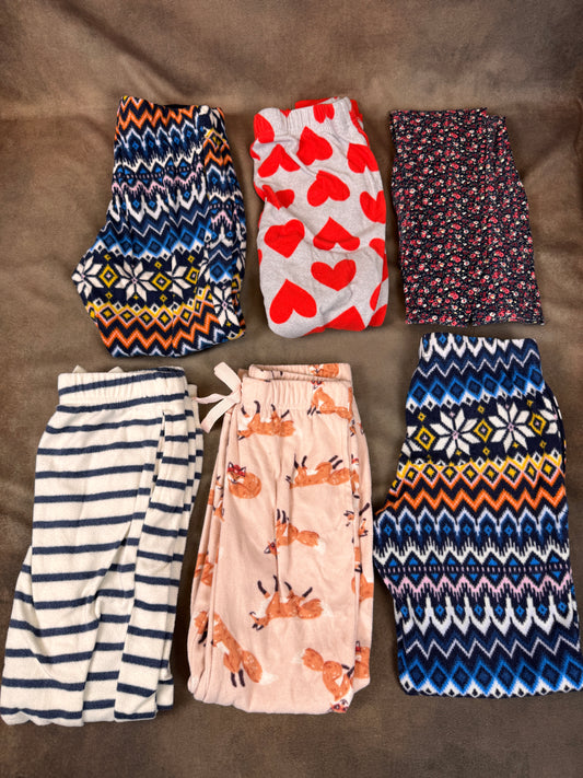Six pairs of folded children's pajama pants arranged in two rows on a brown fabric background. The top row includes patterned sets with geometric and floral motifs, and the bottom row includes a navy-and-white striped pair, a light pink pair with a fox print, and a navy-and-blue patterned pair.